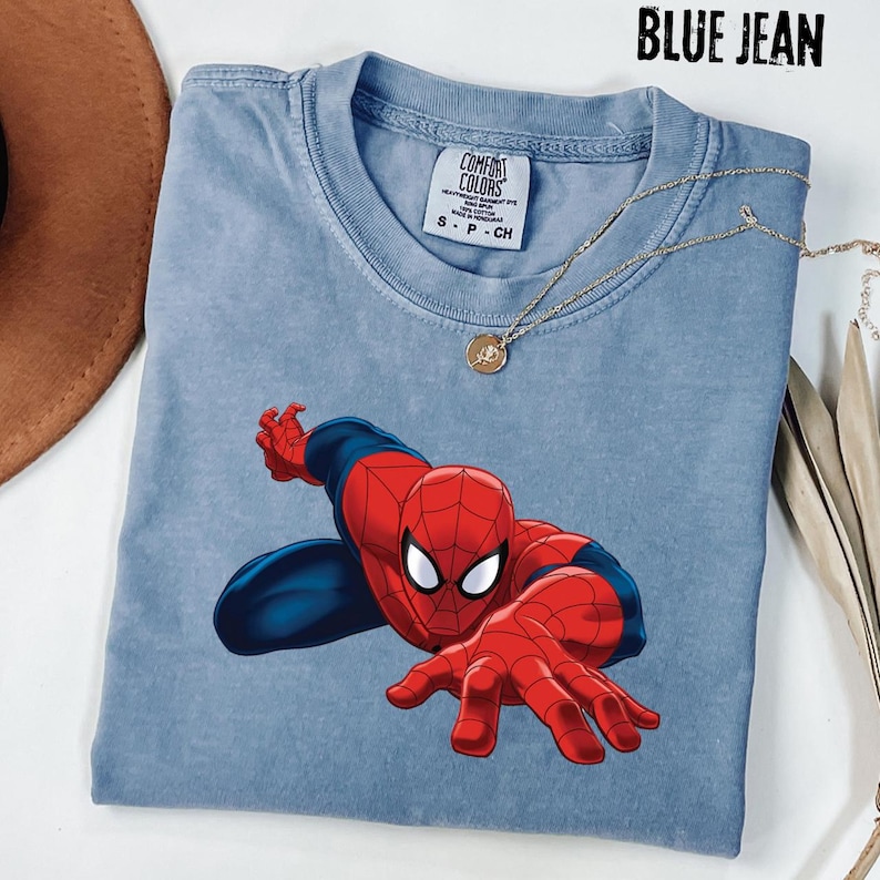 May include: A blue jean colored short-sleeve t-shirt featuring a graphic of the character Spider-Man in a crouching pose with red and blue colors. The shirt has a Comfort Colors brand label on the inside collar and a crew neckline design.