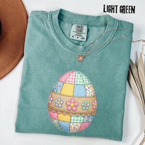 Puede incluir: Camiseta Comfort Colors verde claro con un diseño de huevo de Pascua colorido. El huevo está hecho de tela de retazos con patrones florales y de cuadros. Un collar plateado con un colgante está drapeado sobre la camisa.