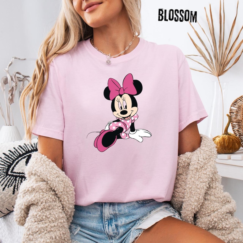 May include: A light pink t-shirt featuring a Minnie Mouse graphic. The cartoon character is depicted in a pink polka-dotted dress and bow. The word "BLOSSOM" is printed in black at the top right.