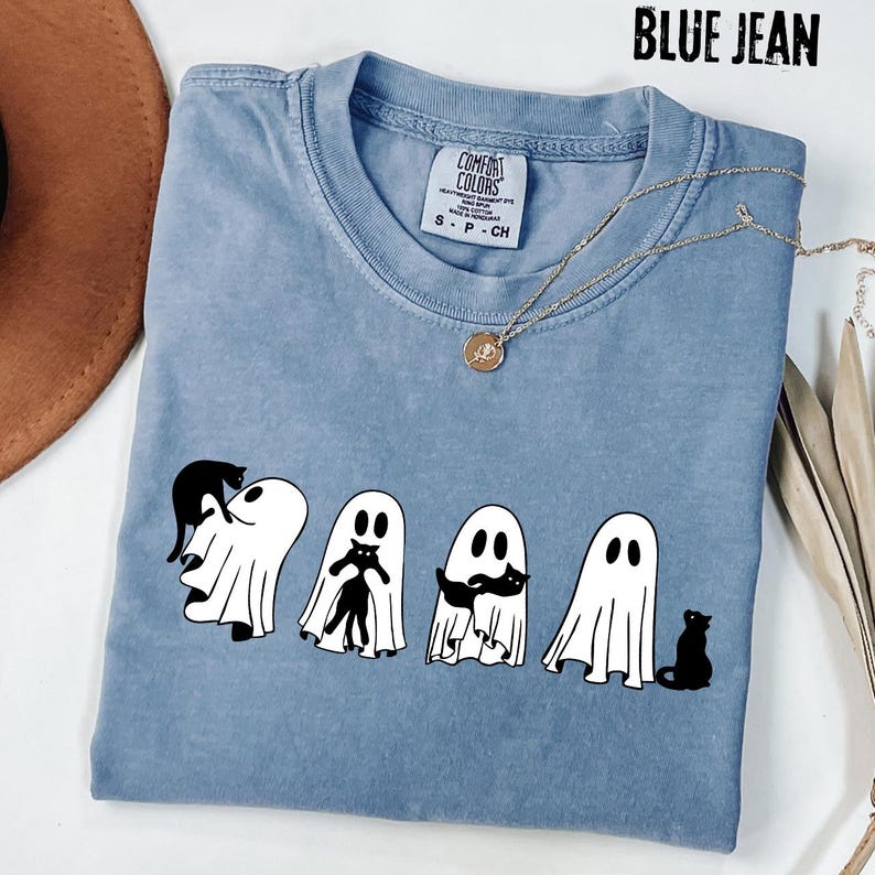 Pu&ograve; includere: Una t-shirt a maniche corte color blu jeans con una grafica raffigurante quattro fantasmi bianchi e gatti neri. La maglietta presenta uno scollo rotondo e un'etichetta del marchio Comfort Colors all'interno del colletto.