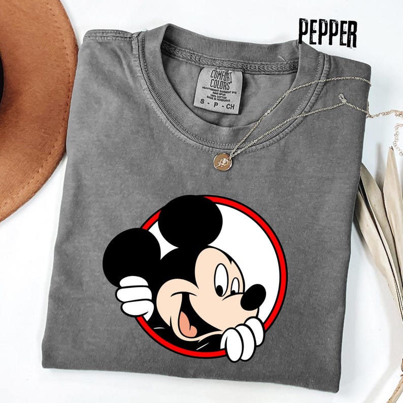 Pu&ograve; includere: Una maglietta grigia Comfort Colors con una grafica di Topolino sorridente all'interno di un cerchio rosso e bianco. La maglietta ha un girocollo e maniche corte. Una collana d'argento con un piccolo ciondolo &egrave; drappeggiata sulla maglietta. La parola "PEPPER" &egrave; stampata in alto.