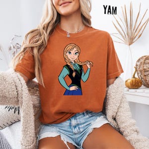 May include: A rust-coloured t-shirt with a cartoon illustration of a character with blonde braids, wearing a blue and black outfit. The word "YAM" is printed in black at the top right. The shirt is a comfortable, casual style.