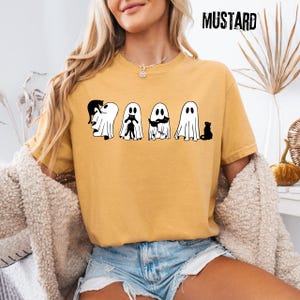 Pu&ograve; includere: Una t-shirt a maniche corte color giallo senape con una stampa grafica di quattro fantasmi bianchi che interagiscono con gatti neri. Il testo MUSTARD &egrave; stampato in nero nell'angolo in alto a destra. La maglietta ha un girocollo.