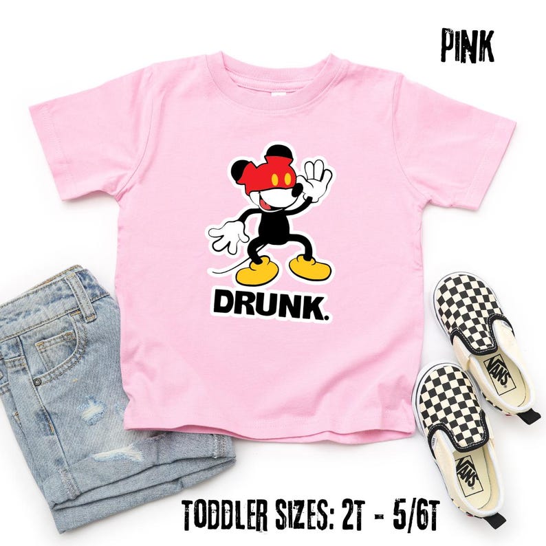 May include: Pink toddler t-shirt featuring a cartoon character with a red hat and the word "DRUNK" in black. The shirt is paired with denim shorts and checkered slip-on shoes. Text at the top reads "PINK" and at the bottom "TODDLER SIZES: 2T - 5/6T".