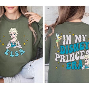 May include: An olive green crewneck sweatshirt featuring a graphic of Elsa on the front with the text ELSA below. The back displays Tinker Bell and Elsa with the text IN MY DISNEY PRINCESS ERA in large blue and white wavy lettering.