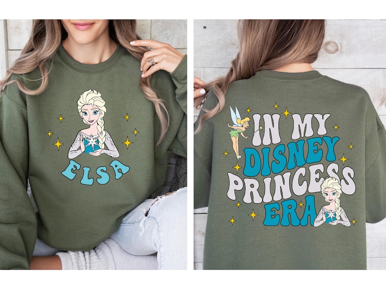 May include: An olive green crewneck sweatshirt featuring a graphic of Elsa on the front with the text ELSA below. The back displays Tinker Bell and Elsa with the text IN MY DISNEY PRINCESS ERA in large blue and white wavy lettering.