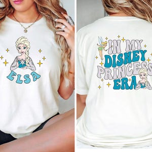May include: A cream-colored t-shirt featuring a graphic of Elsa on the front with the text "ELSA" in blue. The back displays Tinker Bell and Elsa with the text "IN MY DISNEY PRINCESS ERA" in blue and grey, surrounded by yellow star accents.