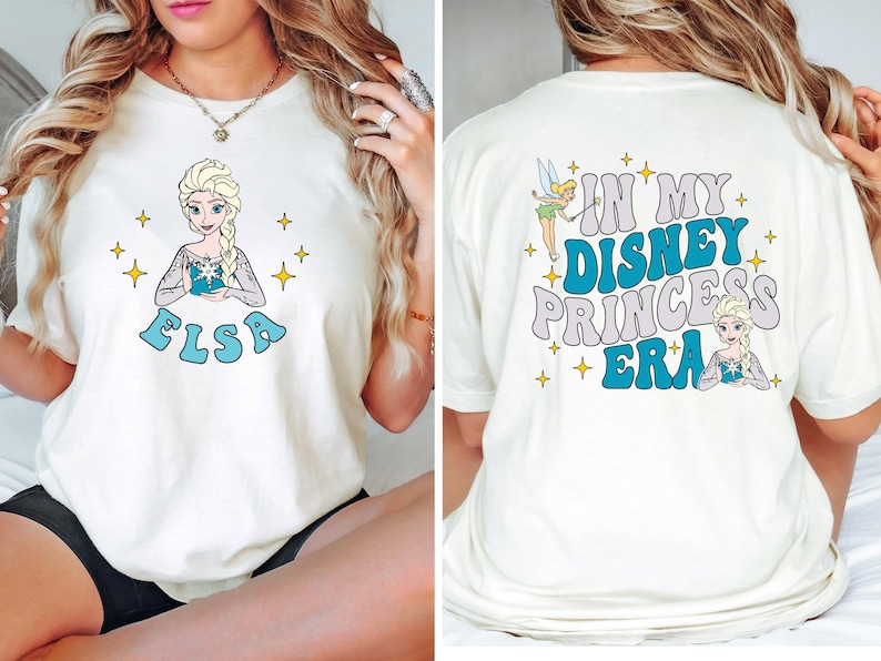 May include: A cream-colored t-shirt featuring a graphic of Elsa on the front with the text "ELSA" in blue. The back displays Tinker Bell and Elsa with the text "IN MY DISNEY PRINCESS ERA" in blue and grey, surrounded by yellow star accents.