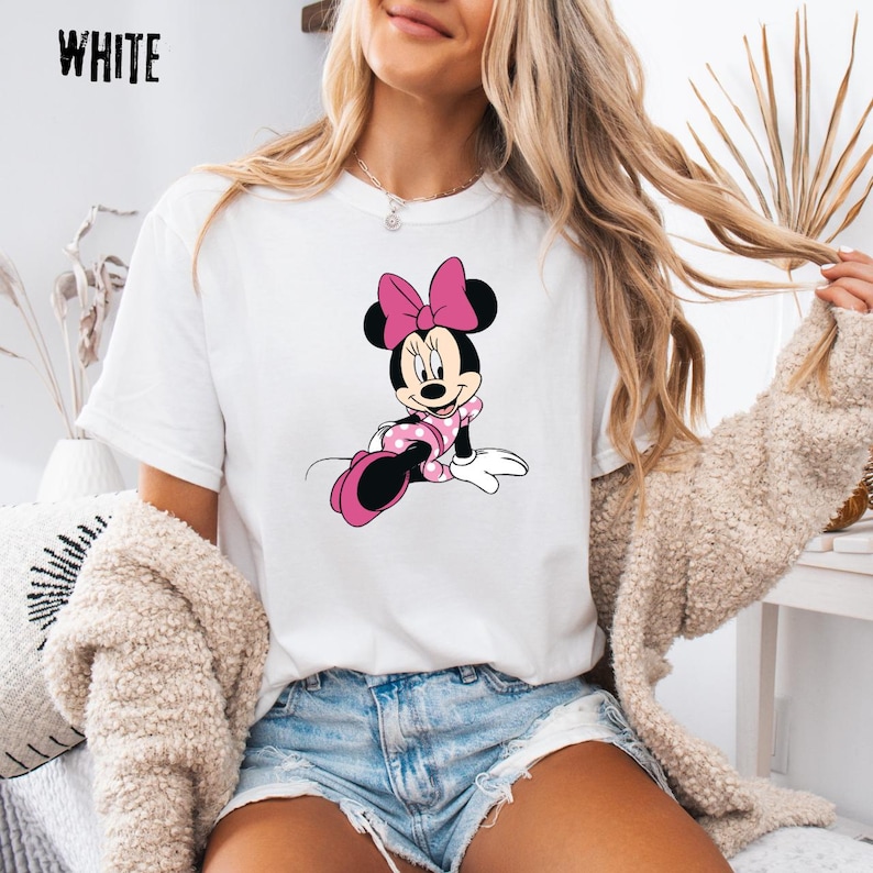 May include: White t-shirt featuring a Minnie Mouse graphic. The cartoon character is depicted in a pink polka-dot dress and bow. The word "WHITE" is printed in black at the top left corner of the shirt.