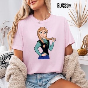 May include: A pale pink t-shirt with a cartoon image of a character with blonde braids, dressed in blue and black. The word "BLOSSOM" is printed in black at the top right corner of the shirt.