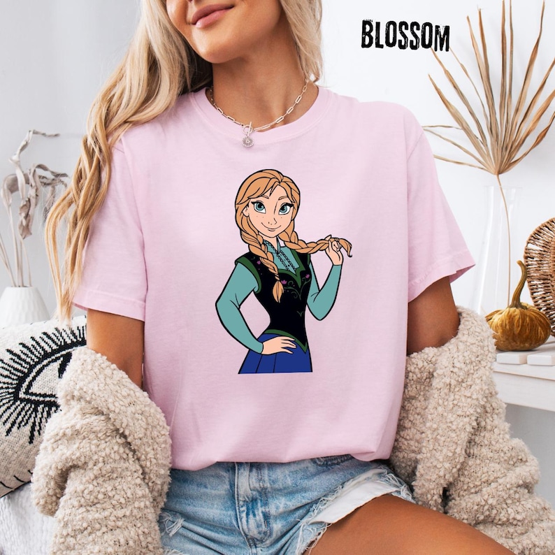 May include: A pale pink t-shirt with a cartoon image of a character with blonde braids, dressed in blue and black. The word "BLOSSOM" is printed in black at the top right corner of the shirt.