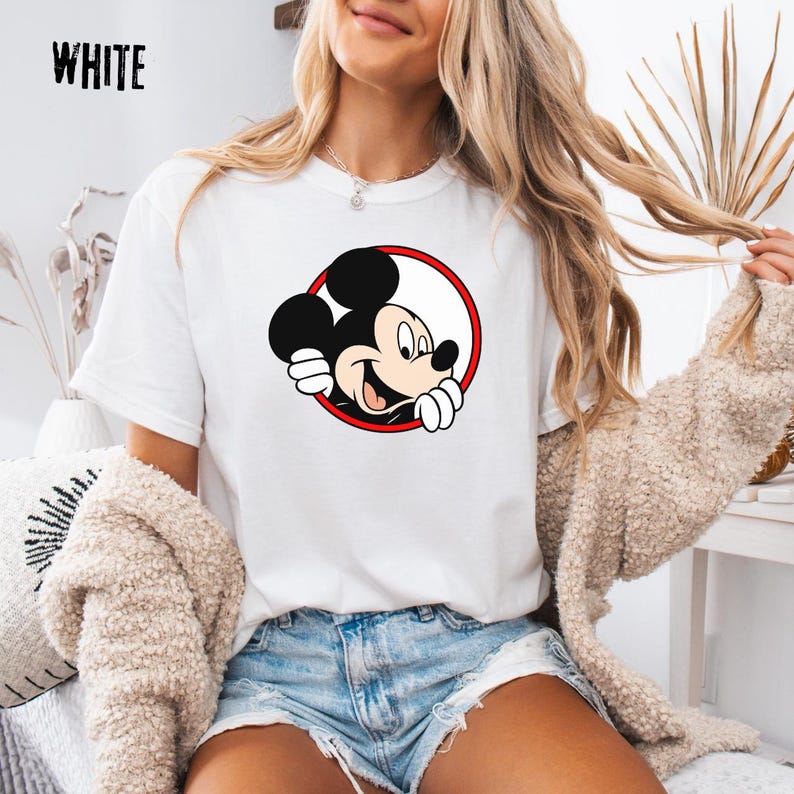 Pu&ograve; includere: T-shirt bianca con grafica di Topolino sorridente all'interno di un cerchio rosso e bianco. La parola "WHITE" &egrave; stampata in nero in alto a sinistra. La maglietta &egrave; in stile casual.