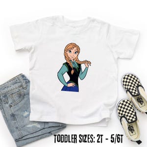 May include: White toddler t-shirt with a cartoon illustration of a character with blonde braids, wearing a blue and black outfit. The text "TODDLER SIZES: 2T - 5/6T" is printed at the bottom. A pair of checkered Vans shoes and denim jeans are also in the image.