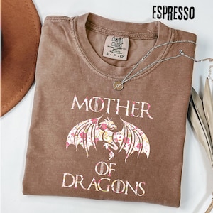 May include: Espresso-coloured t-shirt featuring the text "MOTHER OF DRAGONS" in white. A dragon with floral wings is below the word "MOTHER". A silver necklace with a pendant is draped across the shirt.