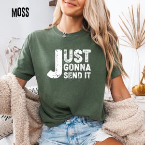 May include: Moss green t-shirt with the text "JUST GONNA SEND IT" in distressed white lettering. The shirt is a short-sleeved crew neck style. The word "MOSS" is printed in black in the upper left corner.