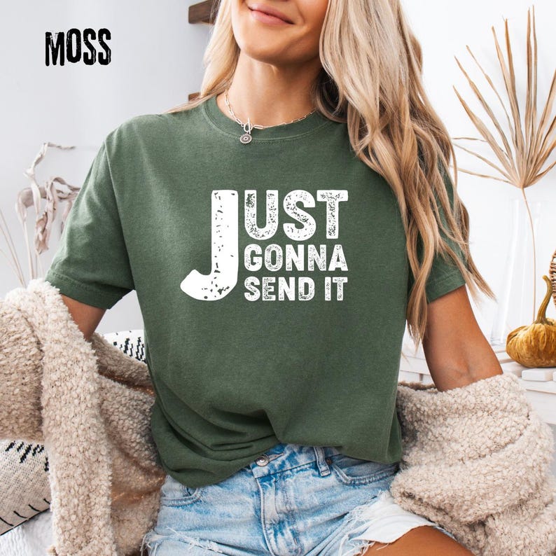 May include: Moss green t-shirt with the text "JUST GONNA SEND IT" in distressed white lettering. The shirt is a short-sleeved crew neck style. The word "MOSS" is printed in black in the upper left corner.