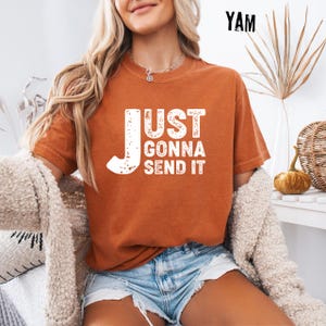 May include: Burnt orange t-shirt with the text "JUST GONNA SEND IT" in white. The shirt is a short-sleeved, crew neck style. The image also shows denim shorts and a cream-colored cardigan.
