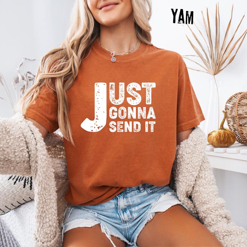 May include: Burnt orange t-shirt with the text "JUST GONNA SEND IT" in white. The shirt is a short-sleeved, crew neck style. The image also shows denim shorts and a cream-colored cardigan.
