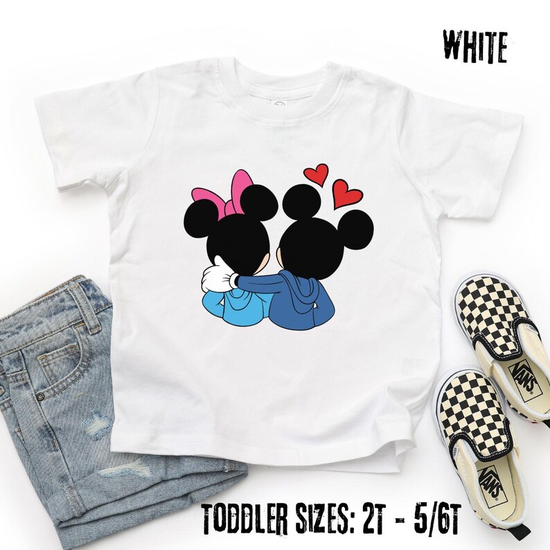 May include: White toddler t-shirt featuring a cartoon illustration of two hugging mouse characters with heart accents. The shirt has the word "WHITE" printed at the top and "TODDLER SIZES: 2T - 5/6T" at the bottom. A pair of black and white checkered Vans shoes and a pair of blue jeans are next to the shirt.