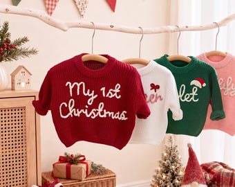 May include: A row of four long-sleeved knit sweaters hanging on a branch. The first is red with white text reading My 1st Christmas. The second is white with red text and a bow. The third is green with white text and a Santa hat. The fourth is pink.