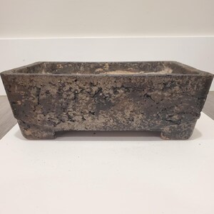 Puede incluir: Una maceta rectangular de color marrón oscuro para bonsái con una superficie texturizada. La maceta tiene una forma ligeramente acampanada y se apoya en cuatro pequeños pies. La estética general es rústica y terrosa, adecuada para plantas pequeñas o bonsáis.