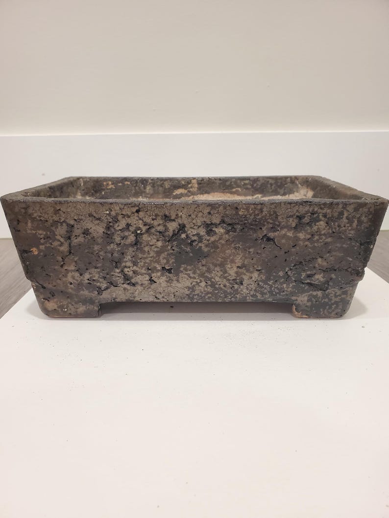 K&ouml;nnte beinhalten: Ein rechteckiger, dunkelbrauner Bonsai-Topf mit strukturierter Oberfl&auml;che. Der Topf hat eine leicht ausgestellte Form und steht auf vier kleinen F&uuml;&szlig;en. Die Gesamt&auml;sthetik ist rustikal und erdig, geeignet f&uuml;r kleine Pflanzen oder Bonsai-B&auml;ume.