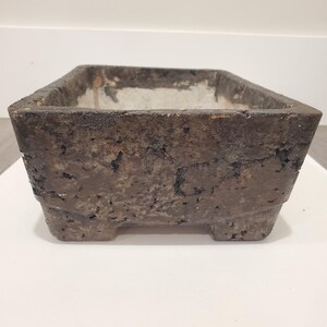 K&ouml;nnte beinhalten: Ein rechteckiger, verwitterter, dunkelbrauner Pflanzk&uuml;bel mit rauer, strukturierter Oberfl&auml;che. Der Pflanzk&uuml;bel hat eine quadratische Form mit einem etwas helleren Innenraum und steht auf vier kleinen F&uuml;&szlig;en. Die Gesamt&auml;sthetik ist rustikal und gealtert.
