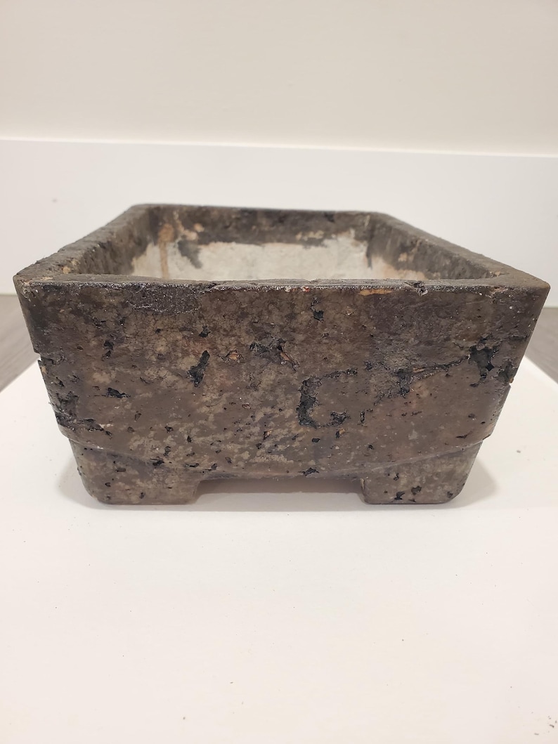 K&ouml;nnte beinhalten: Ein rechteckiger, verwitterter, dunkelbrauner Pflanzk&uuml;bel mit rauer, strukturierter Oberfl&auml;che. Der Pflanzk&uuml;bel hat eine quadratische Form mit einem etwas helleren Innenraum und steht auf vier kleinen F&uuml;&szlig;en. Die Gesamt&auml;sthetik ist rustikal und gealtert.