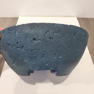 May include: A textured, blue-grey ceramic planter with a wide, oval shape. The planter has a rough, porous surface and small feet. The interior is not visible. Suitable for indoor or outdoor use.