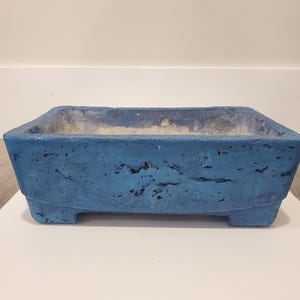 May include: A blue concrete planter, rectangular in shape with a textured finish. The planter has a tapered design and rests on four small feet. The interior has a lighter, weathered look, suitable for plants. Measures approximately 30 cm in length.