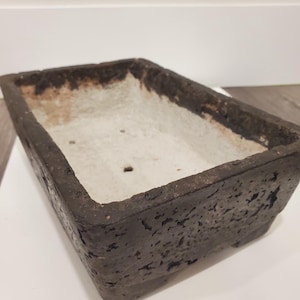 K&ouml;nnte beinhalten: Rechteckiger, dunkelbrauner Pflanztopf mit strukturierter Au&szlig;enseite und hellfarbener Innenseite. Der Pflanztopf hat ein verwittertes Aussehen, was darauf hindeutet, dass er aus Stein oder einem &auml;hnlichen Material besteht. Das Design ist schlicht und funktional.