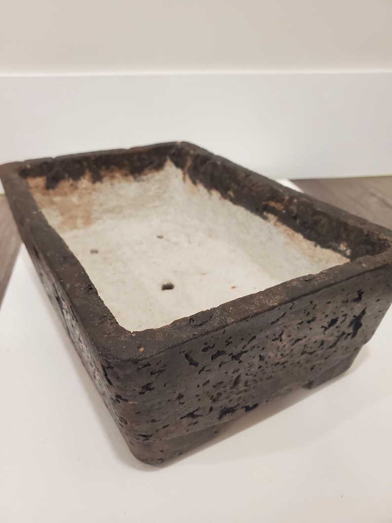 K&ouml;nnte beinhalten: Rechteckiger, dunkelbrauner Pflanztopf mit strukturierter Au&szlig;enseite und hellfarbener Innenseite. Der Pflanztopf hat ein verwittertes Aussehen, was darauf hindeutet, dass er aus Stein oder einem &auml;hnlichen Material besteht. Das Design ist schlicht und funktional.