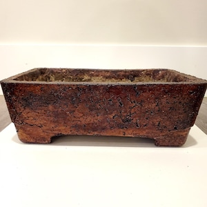 Può includere: Un vaso rettangolare marrone per bonsai con una superficie strutturata. Il vaso ha un aspetto invecchiato, con un esterno marrone scuro e un interno marrone più chiaro. Il vaso è rialzato su quattro piedini.