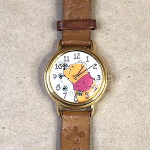 May include: A gold-toned wristwatch with a brown leather band. The watch face features a cartoon image of Winnie the Pooh in a pink shirt, surrounded by blue flowers. The band has a butterfly design.