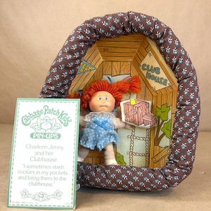 Puede incluir: Una muñeca Cabbage Patch Kids Pin-Ups llamada Charleen Jenny con cabello de lana roja, vistiendo un overol azul con lunares, frente a un recorte de casa club de cartón enmarcado por un arco de tela marrón con un pequeño patrón floral azul.