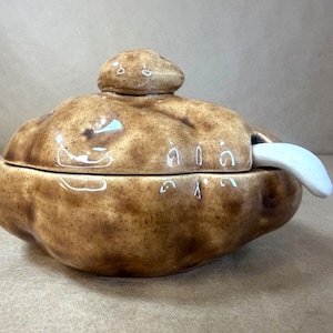 May include: A ceramic lidded container shaped like a potato with a glossy, mottled brown finish. The lid features a small, rounded handle, and a smooth white ceramic spoon is tucked into a side opening, resting against the bowl's rim.