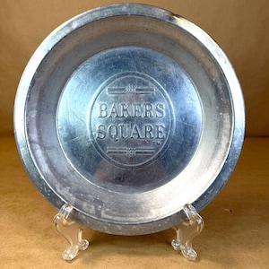 May include: A vintage, round, silver-toned metal pie plate with the embossed text "BAKERS SQUARE" in the center. The plate is displayed on a clear, decorative stand, showcasing its design. The plate has a slightly worn appearance, suggesting age and use.