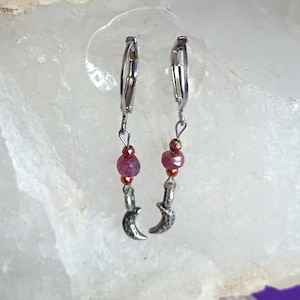 May include: A pair of silver-tone dangle earrings. Each earring features a small, dark crescent moon charm, a red bead, and a smaller copper-colored bead. The earrings are displayed on a large, white, crystalline rock against a purple background.