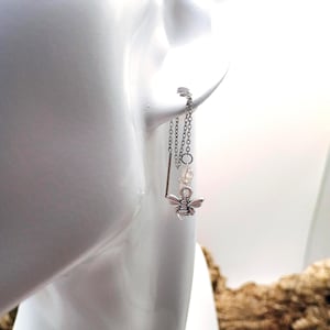 May include: A silver-toned threader earring featuring a delicate chain that passes through the earlobe. The earring includes a small, detailed bee charm and a clear, faceted bead accent, finished with a thin, straight metal bar at the end.