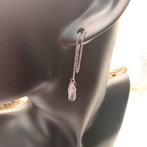May include: A silver-toned chain threader earring featuring a small, clear, raw crystal point pendant wrapped in thin silver wire. The earring is designed to be pulled through the earlobe, with the delicate metal chain hanging down.