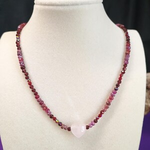 May include: A beaded necklace with a heart-shaped pendant. The necklace features small, faceted beads in shades of red, pink, and purple. The heart pendant is a pale pink color. The necklace is displayed on a white mannequin bust.