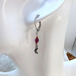 May include: A single dangle earring with a silver-tone lever back. The earring features a cluster of red beads and a small, silver-tone crescent moon charm. The earring is designed to be worn on the ear.