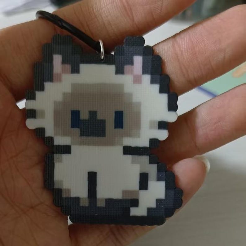 May include: A pixelated cat pendant with a black cord. The pendant features a Siamese cat design with a white body, gray face, and blue eyes. The cat is in a sitting position. The pendant is held in a hand.