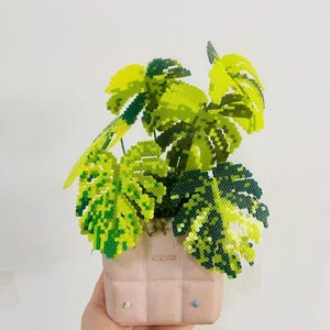 Pode incluir: Uma escultura de planta decorativa feita de pequenas contas de plástico fundidas em tons de verde-limão, amarelo e verde-floresta escuro, imitando a forma de uma planta monstera. A planta está num vaso de cerâmica quadrado de cor rosa pálido.