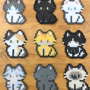 May include: Nine pixelated cat designs in various colors, including black, white, gray, orange, and calico. Each cat is in a sitting position, with a simple, blocky design. The cats are arranged on a wooden surface.