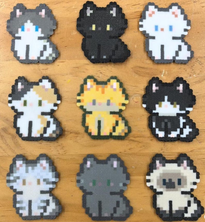 May include: Nine pixelated cat designs in various colors, including black, white, gray, orange, and calico. Each cat is in a sitting position, with a simple, blocky design. The cats are arranged on a wooden surface.
