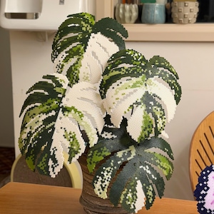 Peut inclure: Une composition végétale décorative avec de grandes feuilles stylisées dans des tons de vert et de blanc, disposées dans un vase marron texturé. Les feuilles ont un aspect pixélisé, créant une esthétique unique et artisanale.