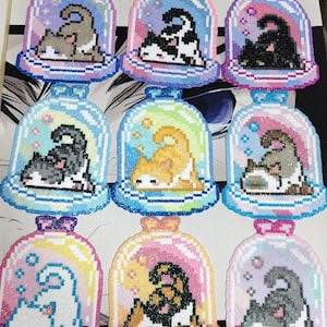 May include: A collection of nine cat-themed ornaments. Each ornament features a pixelated cat inside a glass dome with a colorful background. The cats are in various colors and poses, with glitter accents. The domes have a base and a bow.
