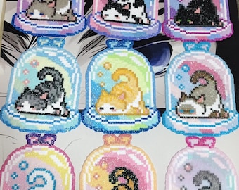 May include: A collection of nine cat-themed ornaments. Each ornament features a pixelated cat inside a glass dome with a colorful background. The cats are in various colors and poses, with glitter accents. The domes have a base and a bow.