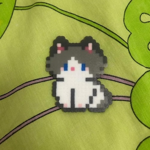 May include: A pixelated cat-shaped pin with a white body, gray head, and pink accents. The cat has blue eyes and black details. The pin is set against a green background with a floral pattern.